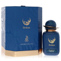 A clear glass bottle with a gold cap and dark amber liquid, standing on a white surface, labeled Arabiyat Prestige Bedour Eau