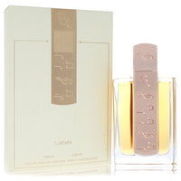 A clear glass bottle with gold accents and a black cap, labeled Lattafa Angham, standing on a white surface by The Fragrance 