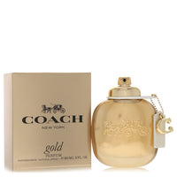 A gold perfume bottle with a sleek design and white label, standing on a wooden surface, labeled Coach Gold Eau De Parfum Spr