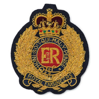 Royal Engineers blazer badge in gold with black enamel, featuring a crossed rifle and pickaxe, mounted on a dark blue backgro