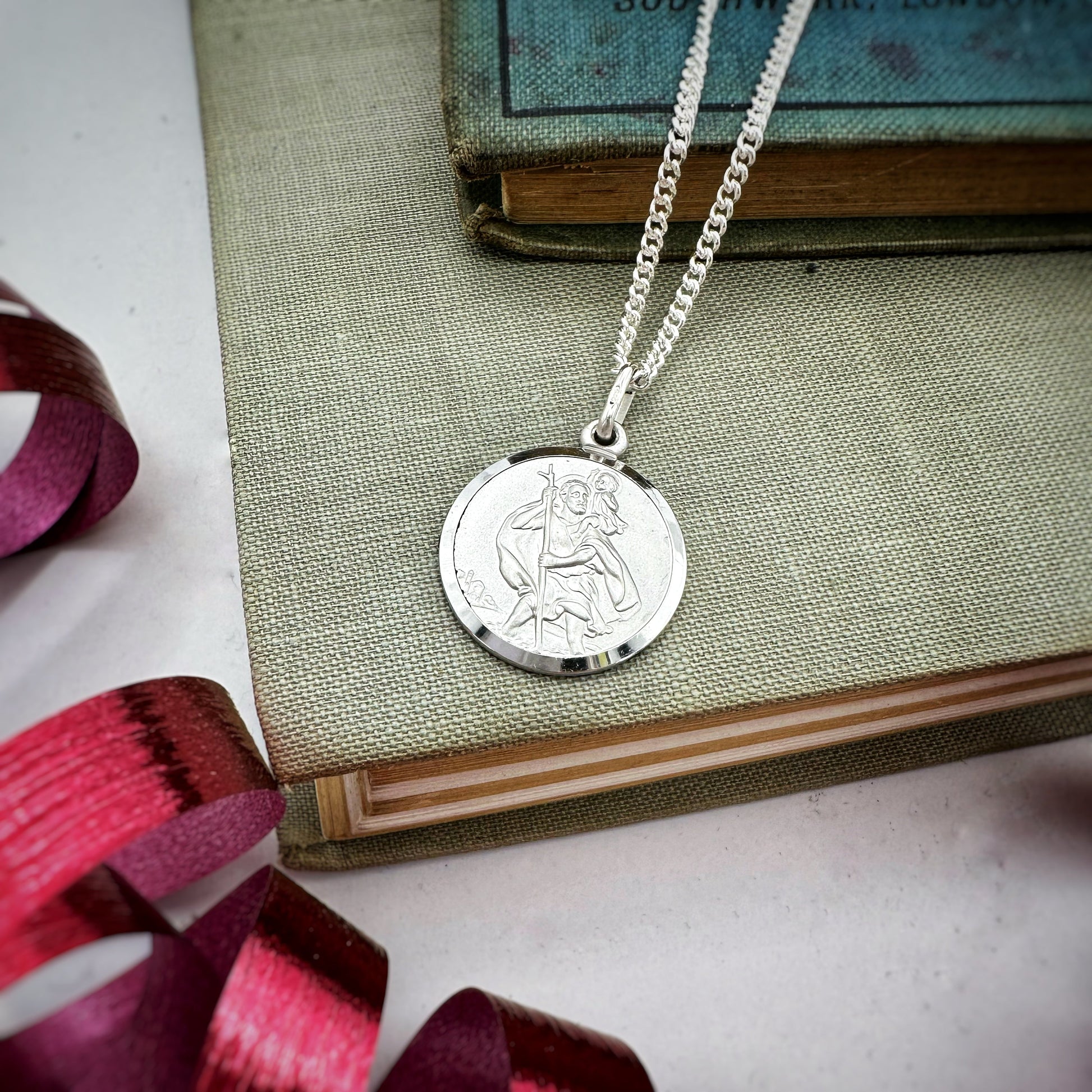 Sterling silver Classic St Christopher Necklace on a green book, showcasing a detailed pendant by Scarlett Jewellery.