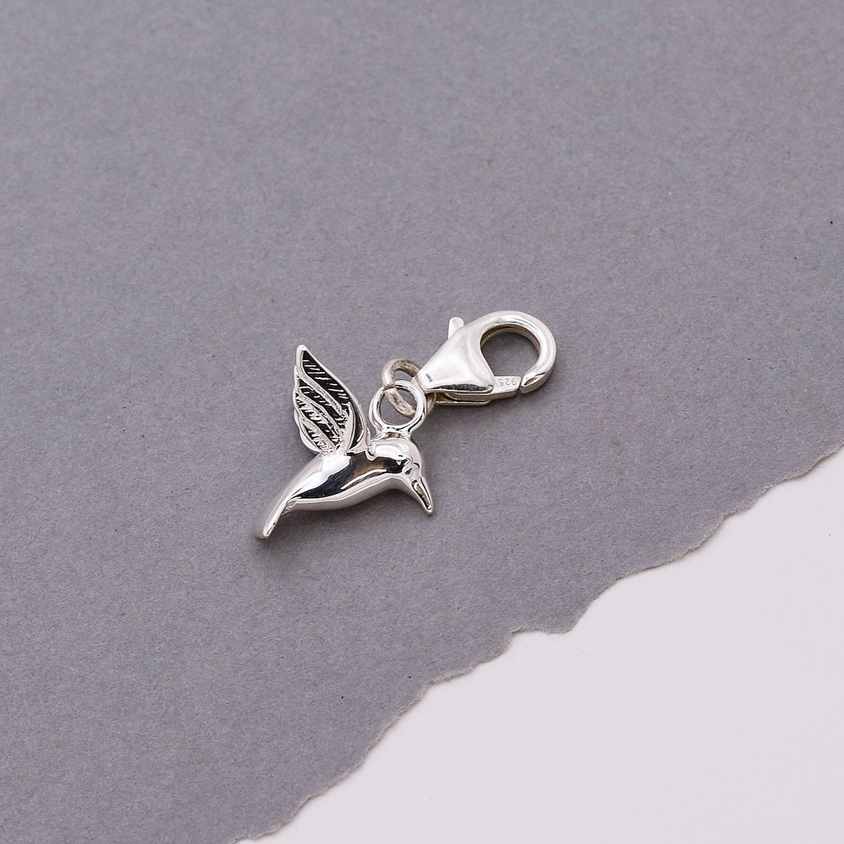 A silver hummingbird charm shaped like a delicate winged bird, displayed on a soft white background by Scarlett Jewellery.