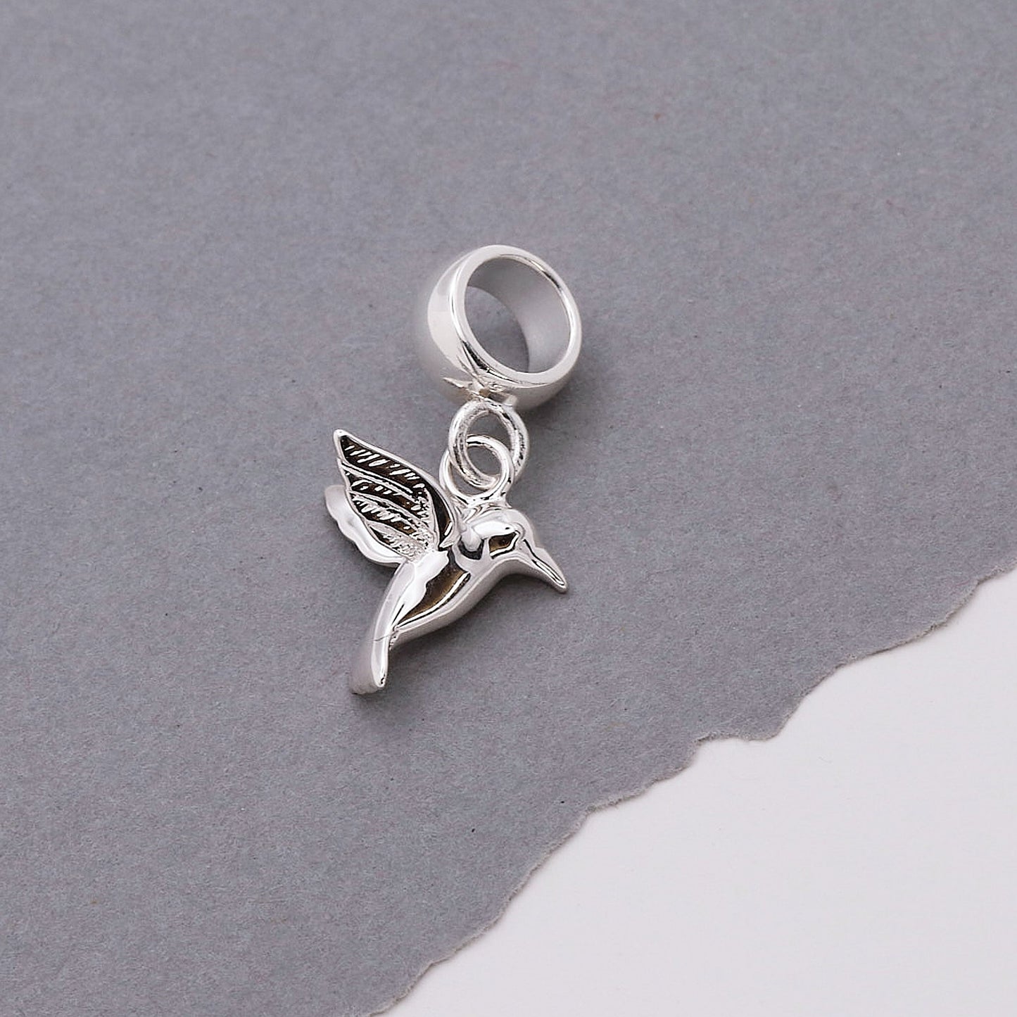 A silver hummingbird charm shaped like a delicate winged bird, featured on a soft white background by Scarlett Jewellery.