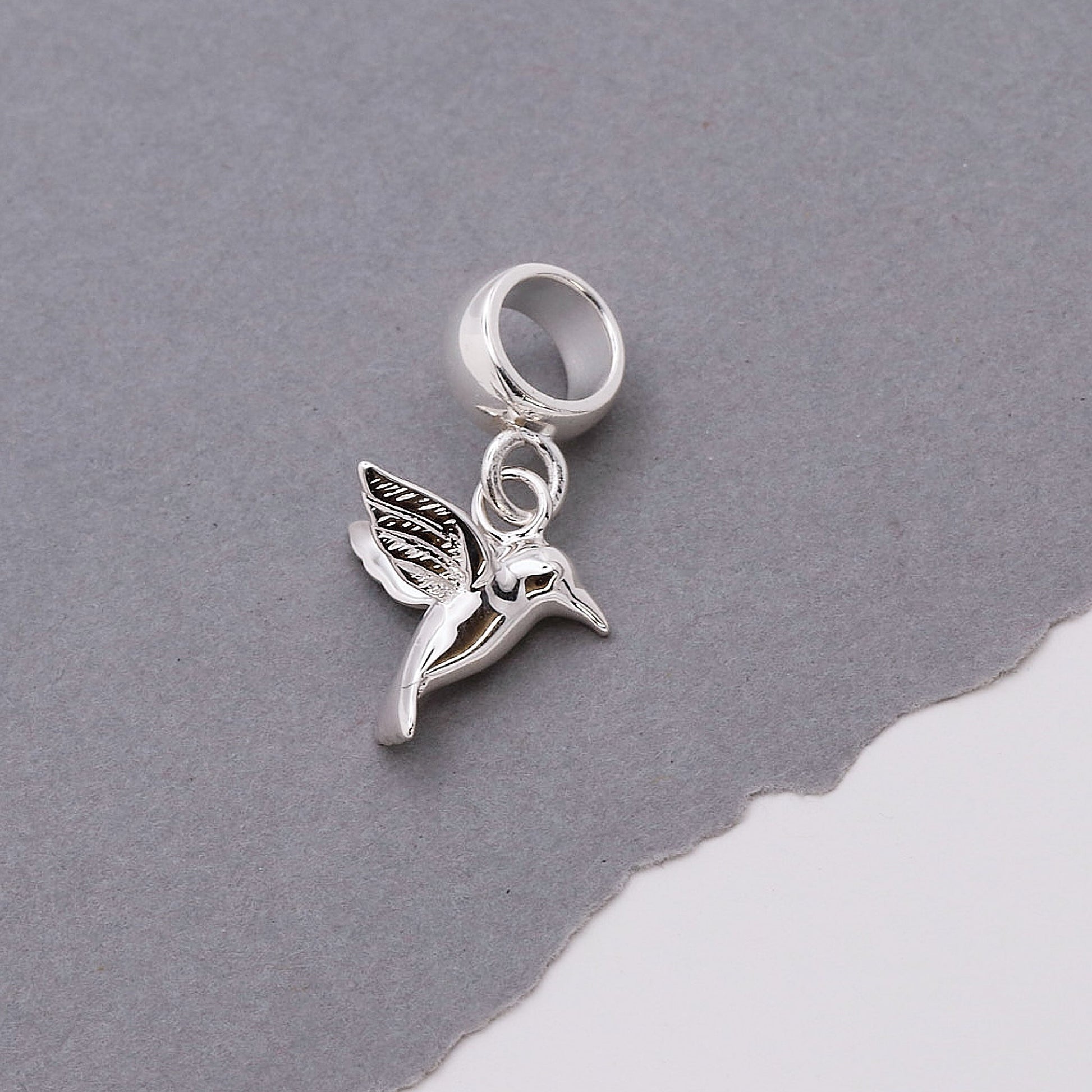 A silver hummingbird charm shaped like a delicate winged bird, featured on a soft white background by Scarlett Jewellery.
