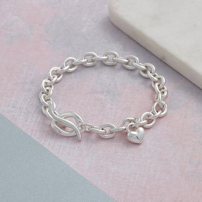 A silver heart charm bracelet with a detailed engraved design, worn on a wrist by Scarlett Jewellery.