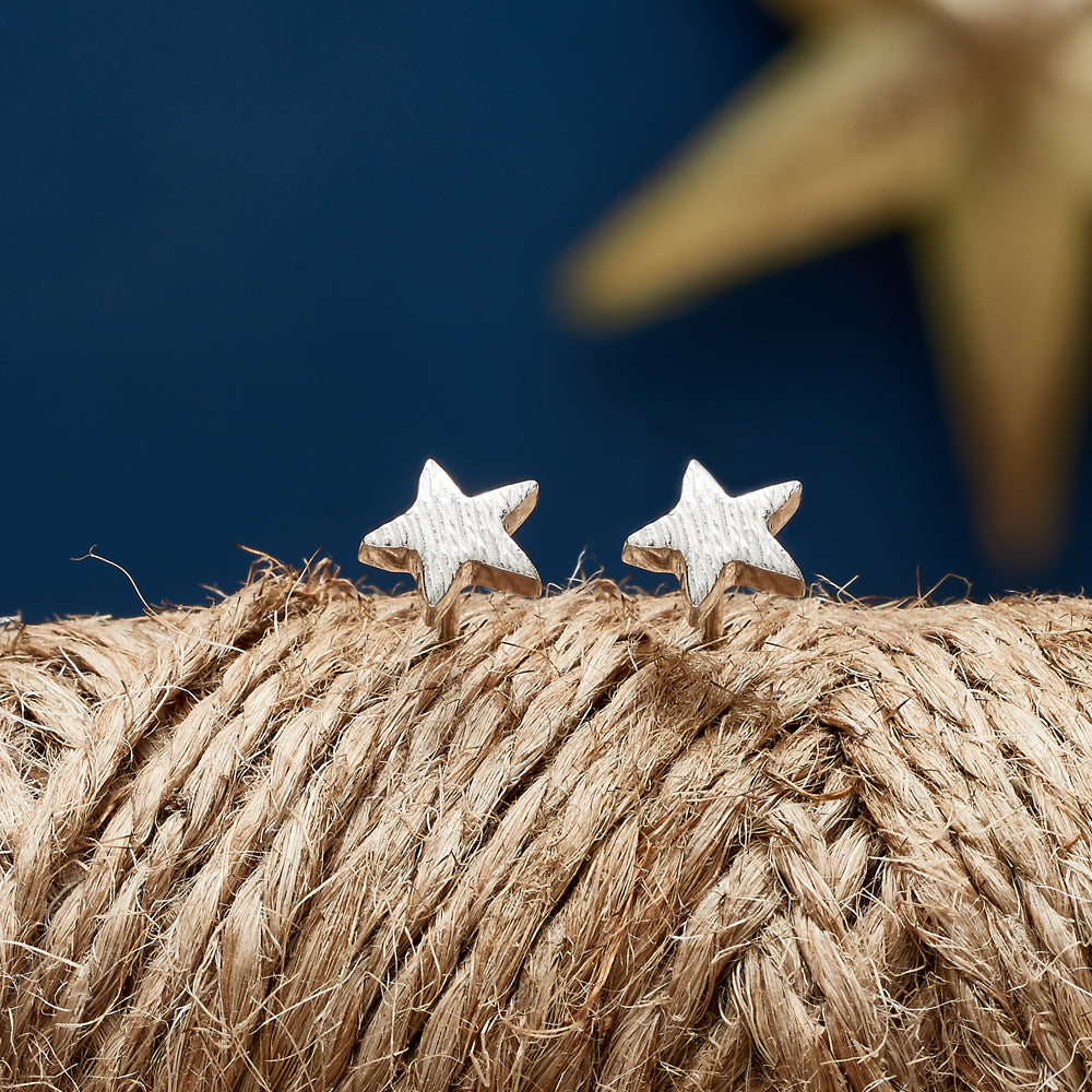 Silver stud earrings with a twinkling star design, styled on a soft pink background by Scarlett Jewellery.