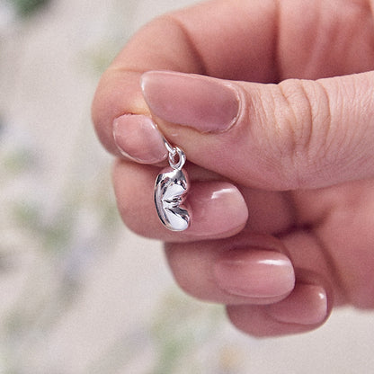 A silver baby bean charm hangs from a chain, detailed and reflective, by Scarlett Jewellery.