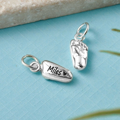 Silver baby charm with personalized engraving, displayed on a soft pink background, by Scarlett Jewellery.