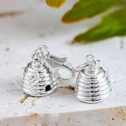 A silver beehive charm with intricate detailing, displayed on a soft pink background, by Scarlett Jewellery.