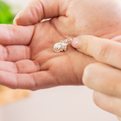 A silver cat charm with intricate detailing, displayed on a soft fabric background, by Scarlett Jewellery.