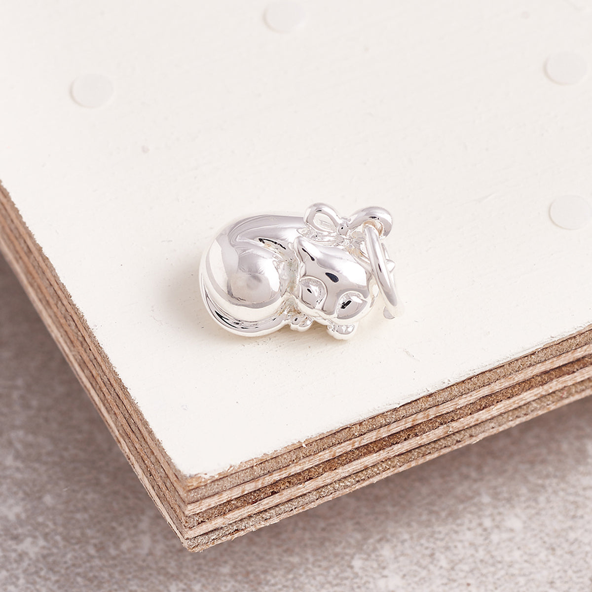 A silver cat charm with intricate detailing, displayed on a soft fabric background, by Scarlett Jewellery.
