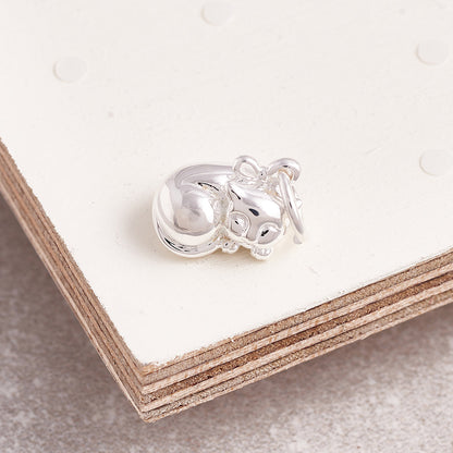 A silver cat charm with intricate detailing, displayed on a soft fabric background, by Scarlett Jewellery.