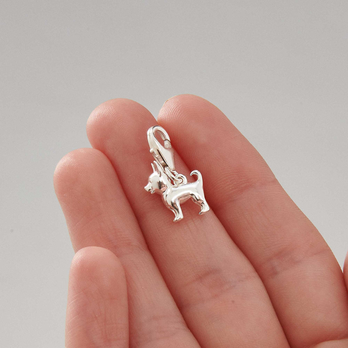 A silver chihuahua dog charm with detailed facial features, displayed on a soft pink background by Scarlett Jewellery.