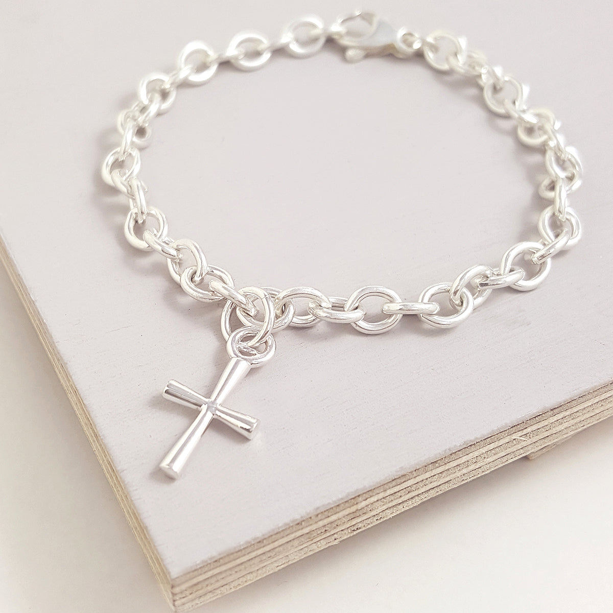 A silver cross charm with intricate detailing, displayed on a soft white background, by Scarlett Jewellery.