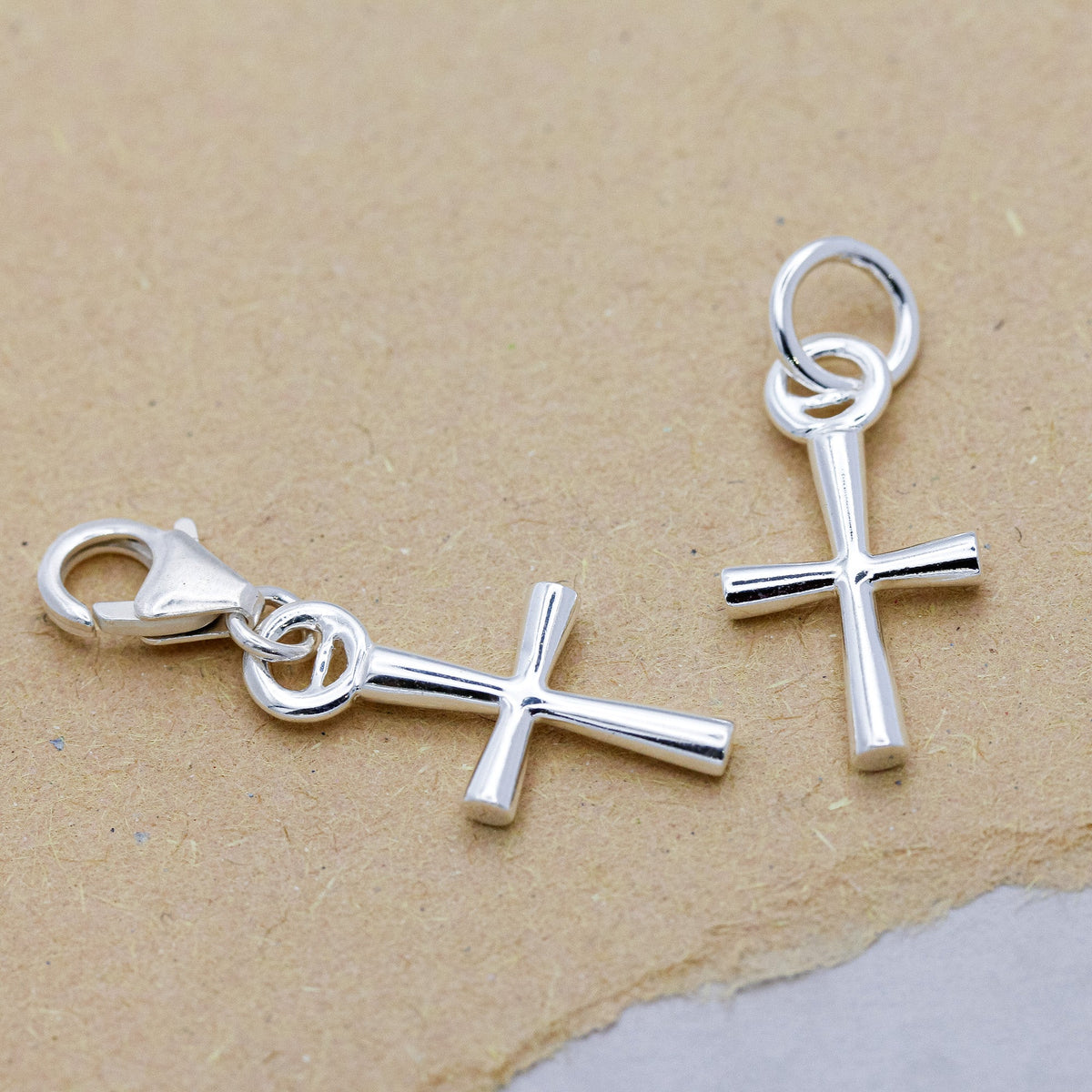 A silver cross charm with intricate detailing, displayed on a soft white background by Scarlett Jewellery.