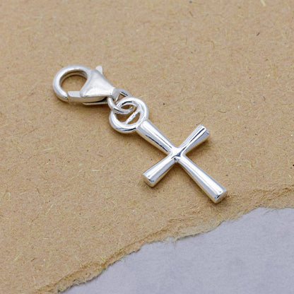 A silver cross charm with intricate detailing, displayed on a soft white background, by Scarlett Jewellery.