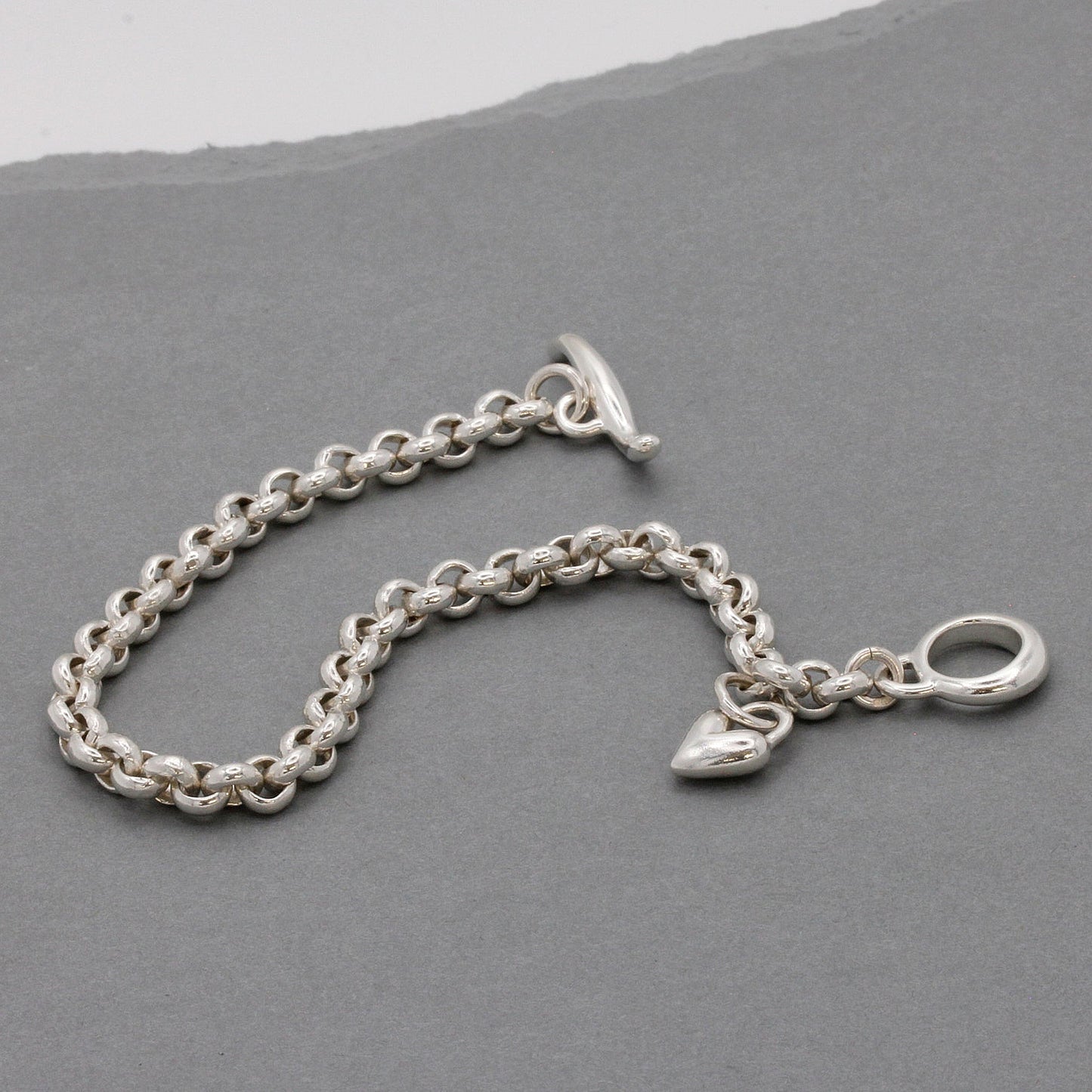 A silver heart chain charm bracelet with a detailed heart pendant, worn on the wrist by Scarlett Jewellery.