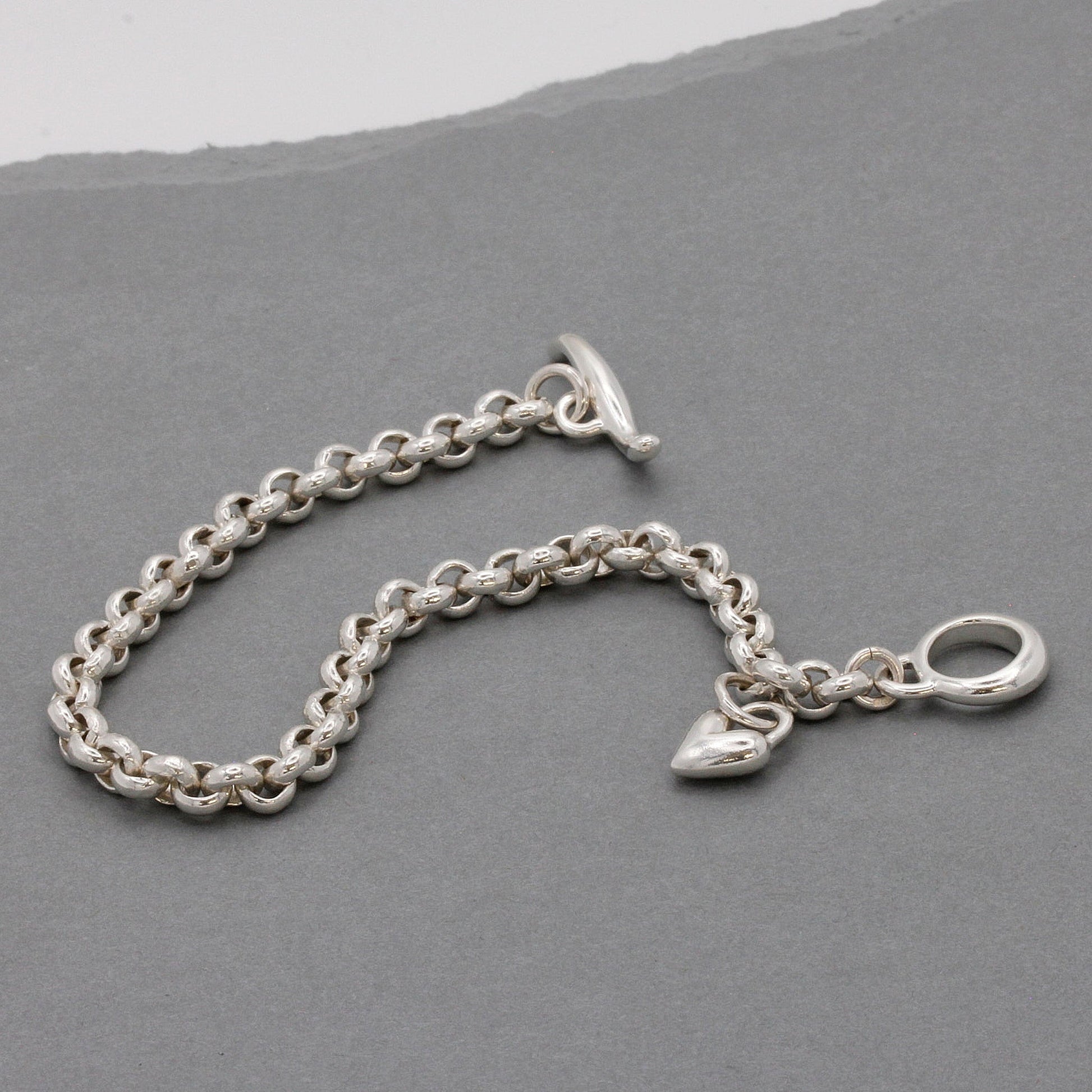 A silver heart chain charm bracelet with a detailed heart pendant, worn on the wrist by Scarlett Jewellery.