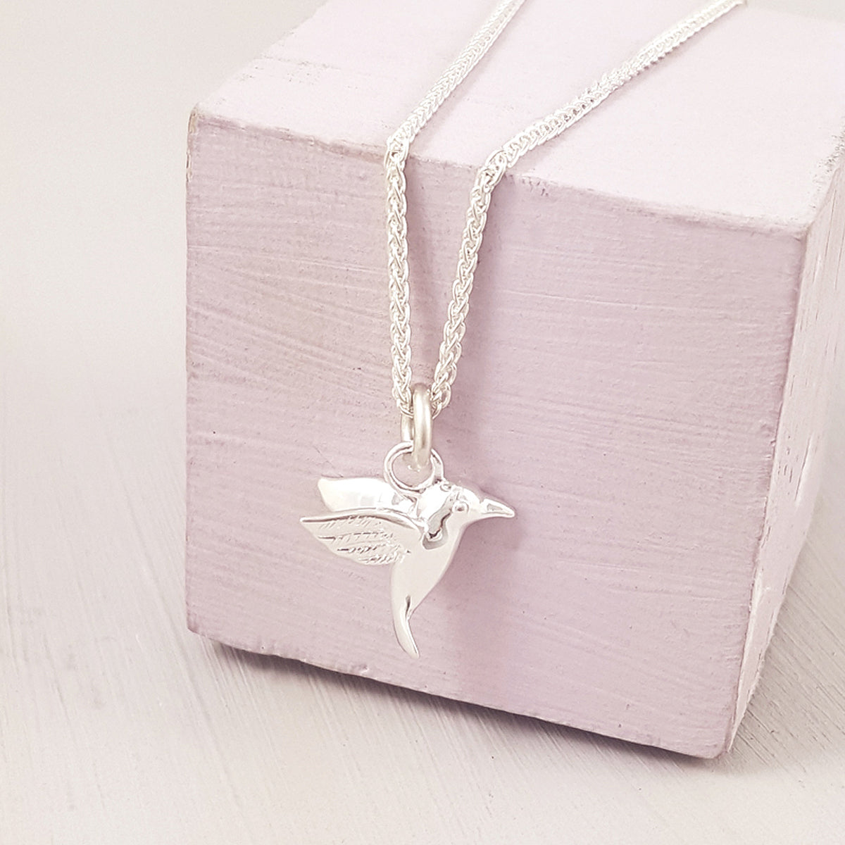 A silver hummingbird charm with detailed wings resting on a velvet background by Scarlett Jewellery.
