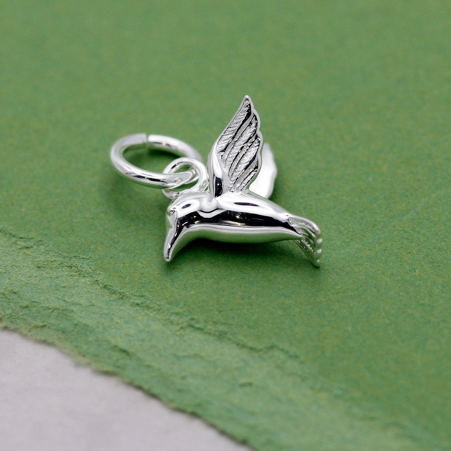 A silver hummingbird charm shaped like a delicate winged bird, displayed on a soft white background by Scarlett Jewellery.
