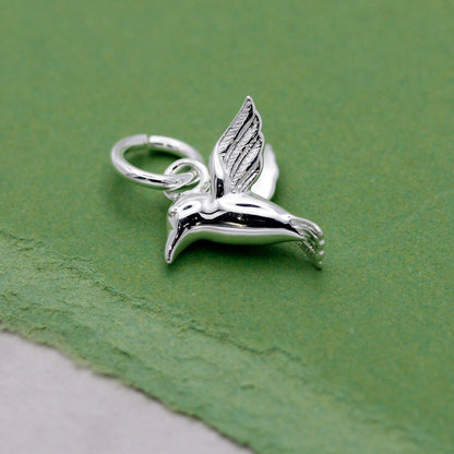 A silver hummingbird charm shaped like a delicate winged bird, displayed on a soft white background by Scarlett Jewellery.