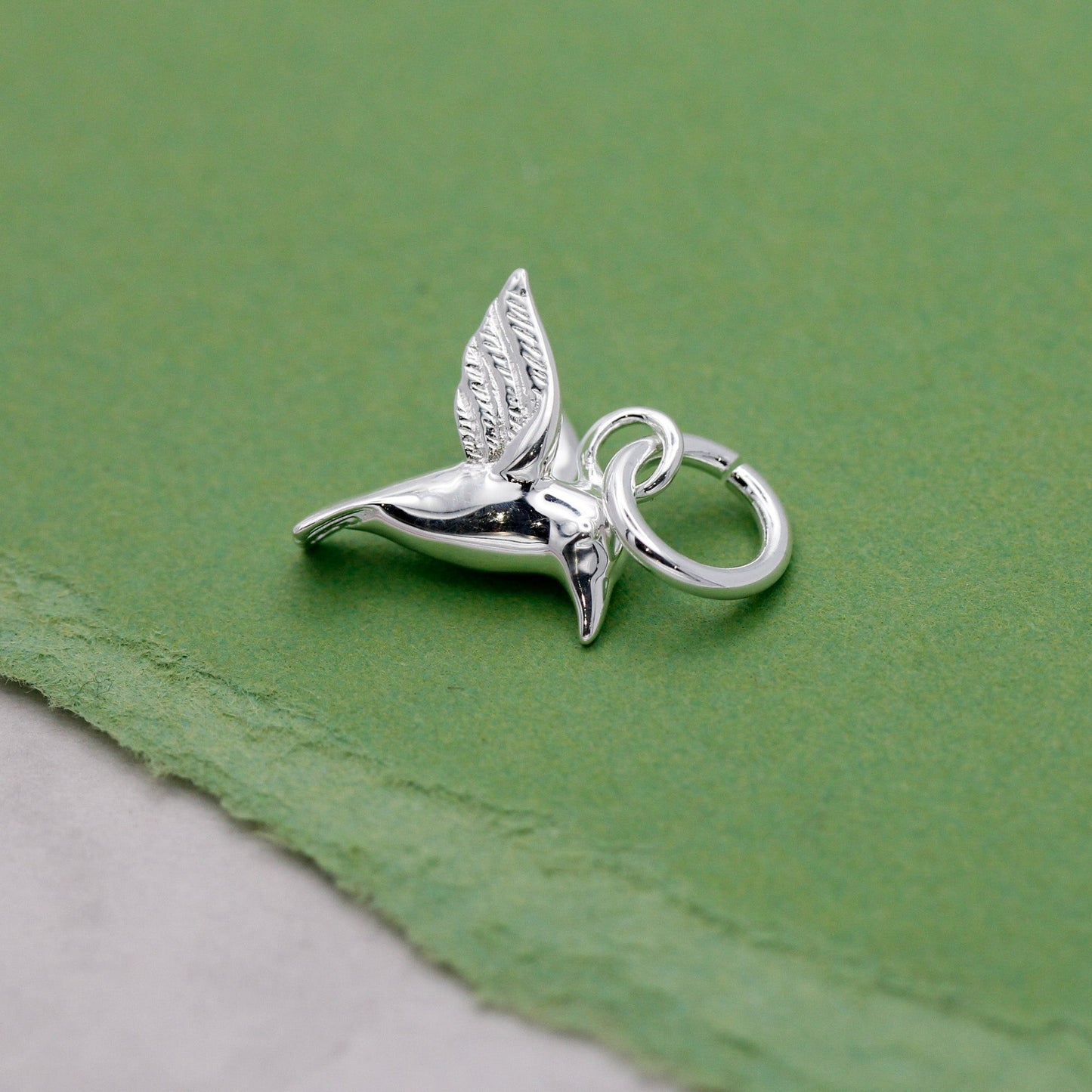A silver hummingbird charm shaped like a delicate winged bird, displayed on a soft white background by Scarlett Jewellery.