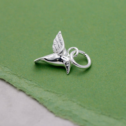 A silver hummingbird charm shaped like a delicate winged bird, displayed on a soft white background by Scarlett Jewellery.