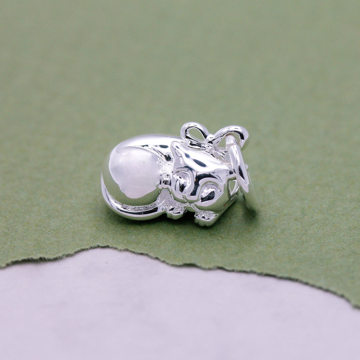 A silver cat charm with intricate detailing, displayed on a soft fabric background, by Scarlett Jewellery.
