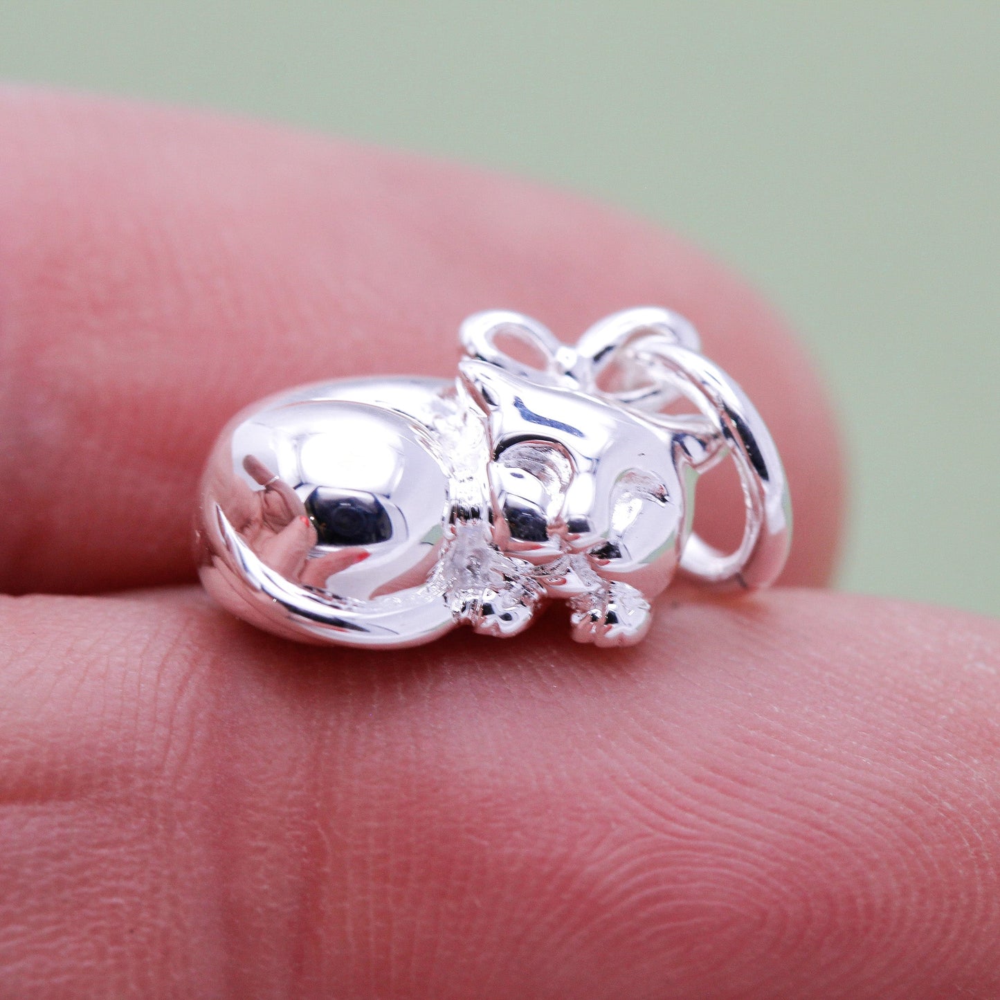 A silver cat charm with intricate detailing, displayed on a soft pink background, part of the Scarlett Jewellery collection.