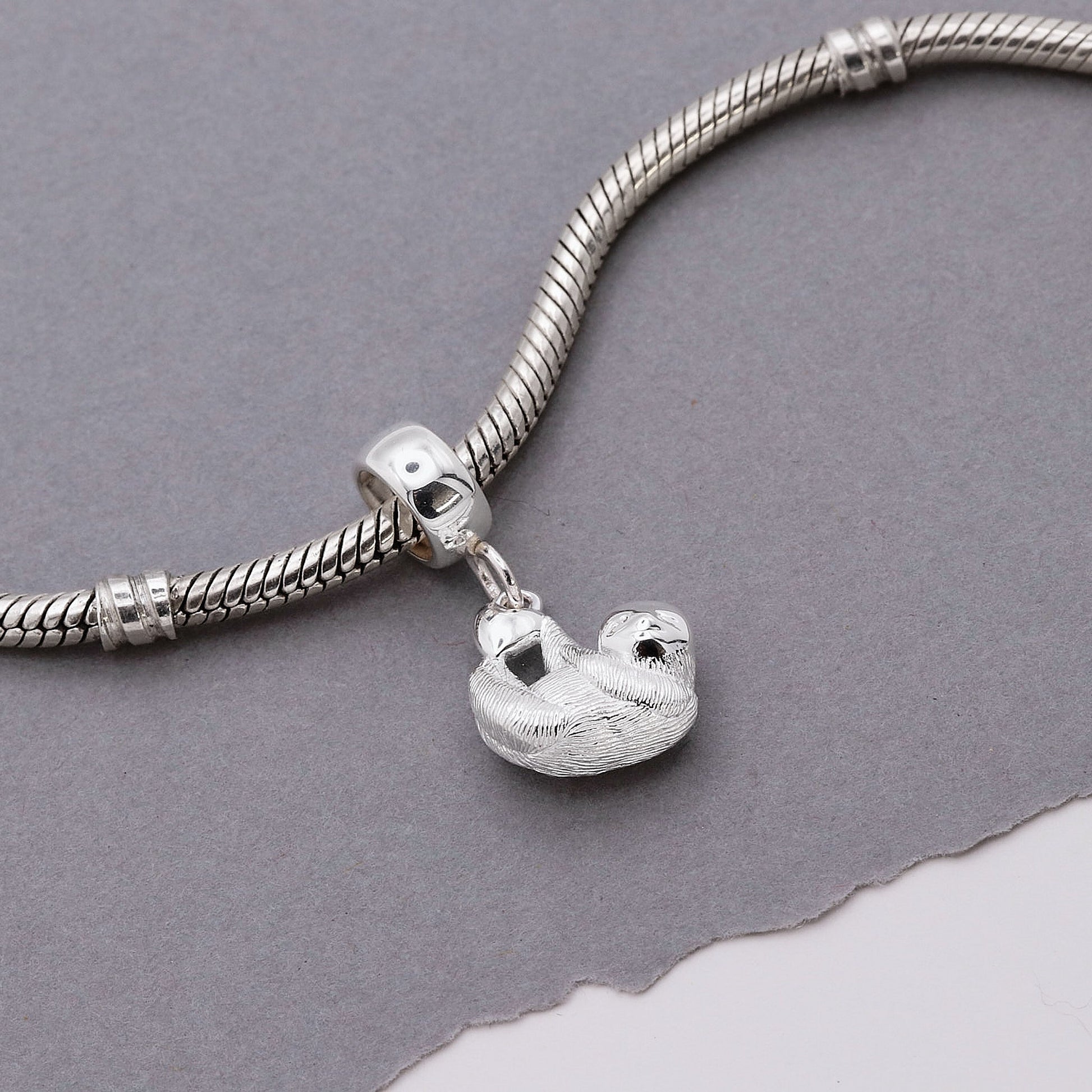 A silver sloth charm shaped like a small pendant, displayed on a soft fabric background by Scarlett Jewellery.