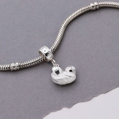 A silver sloth charm shaped like a small pendant, displayed on a soft fabric background by Scarlett Jewellery.