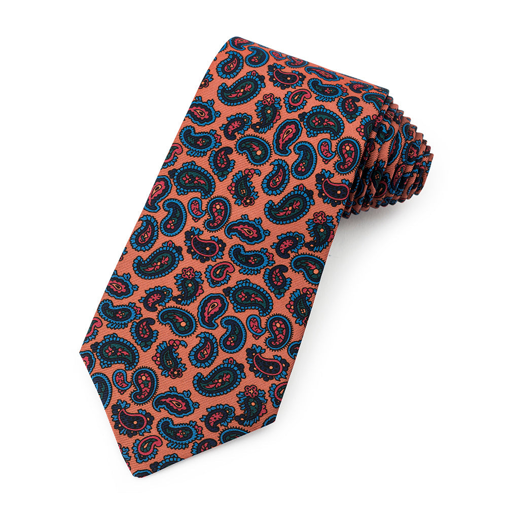 A mustard-colored silk tie featuring a paisley pattern and ancient madder dye, displayed horizontally with brand logo at the 